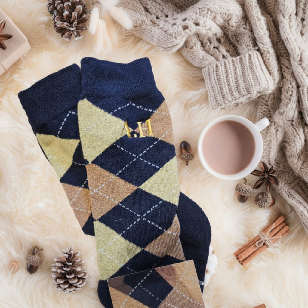 Pair of argyle patterned socks on a soft surface with a cup of hot chocolate and festive decorations.