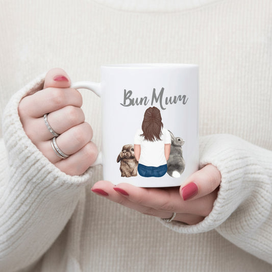 A white ceramic mug with 'Bun Mum' text and illustrations of a woman sitting and two rabbits, held in a person's hands.