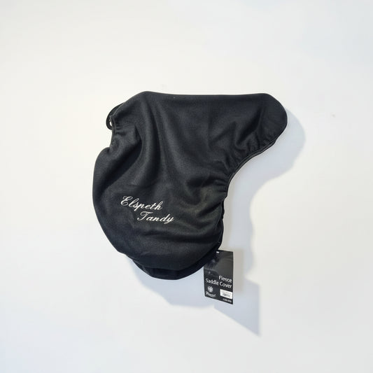 A black fleece horse saddle cover with white embroidered text reading 'Elizabeth' and 'Fandy'. The cover is displayed on a hanger against a wooden background.